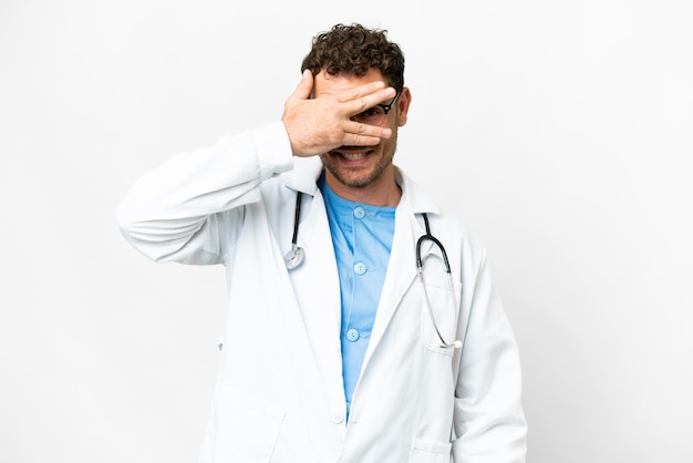 Homme médecin brésilien sur fond blanc isolé couvrant les yeux par les mains et souriant