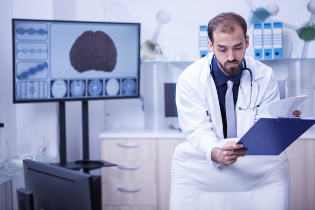 Photo homme médecin dans son cabinet de l'hôpital en regardant ses notes du presse-papiers. cerveau exposé en arrière-plan.