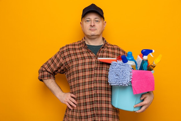 Homme de ménage en chemise à carreaux et casquette tenant un seau avec des outils de nettoyage regardant la caméra avec une expression sérieuse et confiante debout sur fond orange