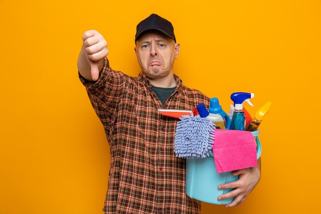 Photo homme de ménage en chemise à carreaux et casquette tenant un seau avec des outils de nettoyage regardant la caméra mécontent montrant les pouces vers le bas debout sur fond orange