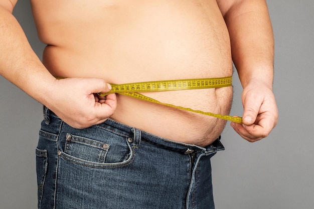 Un homme mesure son gros ventre avec un ruban à mesurer sur fond gris