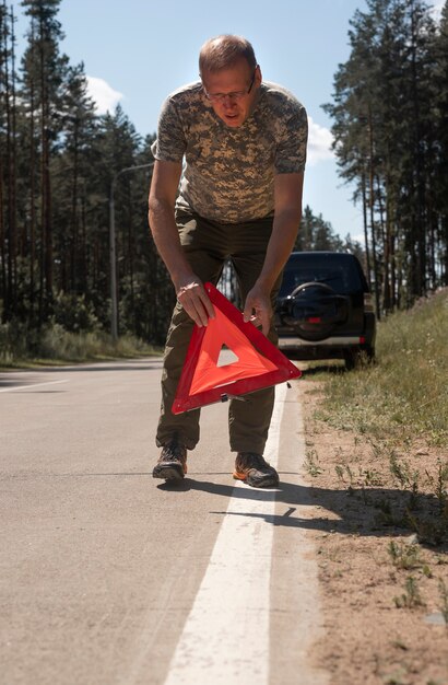 Homme mettant un signe de prudence triangle sur route près d'une voiture cassée au bord de la route