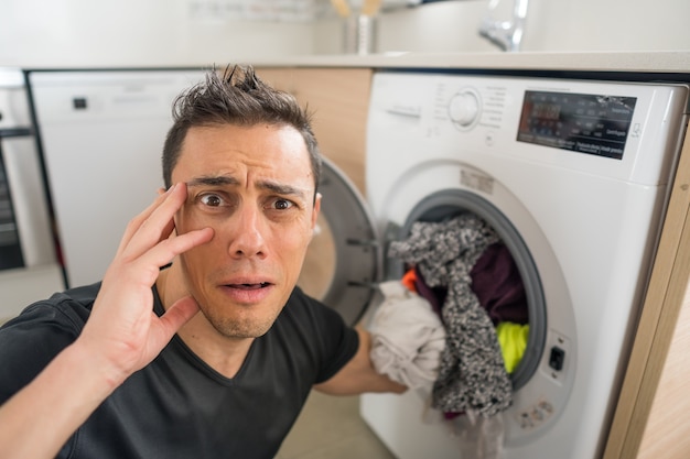 Homme mettant des vêtements dans la machine à laver, dans la cuisine, inquiet car il doit laver beaucoup de vêtements. Fermer.