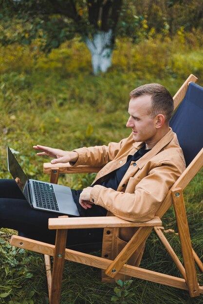 Homme mignon dans une chemise assis sur une chaise à l'extérieur dans le jardin et travaillant sur un ordinateur portable