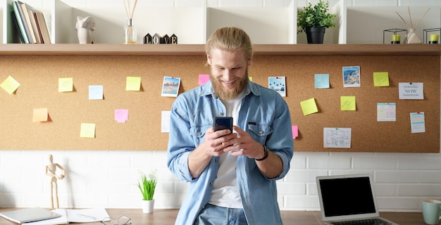 Homme millénaire souriant debout dans un espace de travail à domicile en utilisant un téléphone portable