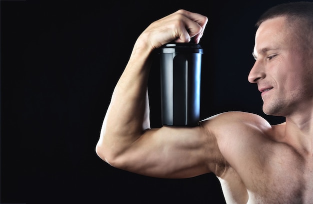 Un homme mince et fort tient sur un biceps un shaker noir avec des protéines. Guy pose sur le fond sombre.