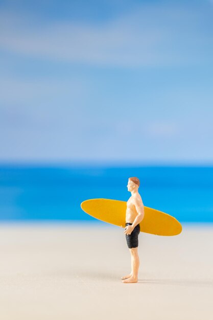 Un homme en miniature en maillot de bain tenant une planche de surf jaune sur la plage.