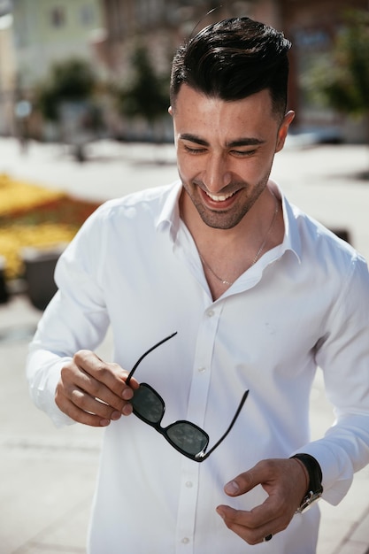 Un homme de mode en chemise blanche se tient dans la rue par une journée ensoleillée pose en regardant des lunettes
