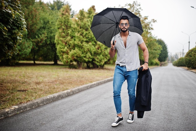 Homme à la mode grand barbe arabe porter sur chemise jeans et lunettes de soleil marchant au parc avec parapluie et manteau à portée de main