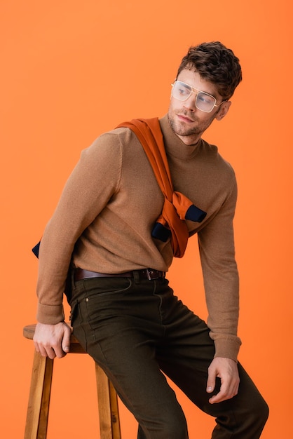 Homme à la mode en tenue d'automne et lunettes assis sur un tabouret en bois isolé sur orange