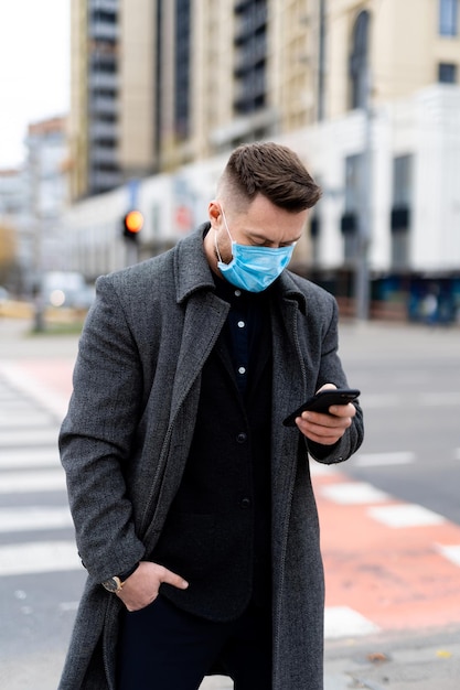 Homme à la mode en vêtements formels portant un masque de sécurité et utilisant un téléphone à l'extérieur. Comportement responsable pendant la pandémie mondiale de covid-19.
