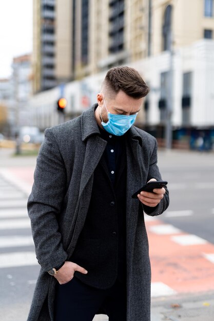 Homme à la mode en vêtements formels portant un masque de sécurité et utilisant un téléphone à l'extérieur. Comportement responsable pendant la pandémie mondiale de covid-19.