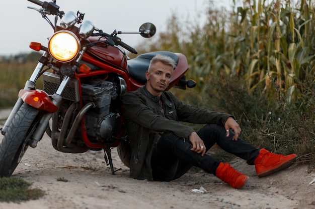 Homme modèle brutal dans une veste kaki militaire de mode avec des baskets élégantes rouges se trouve près d'une moto dans un champ de maïs