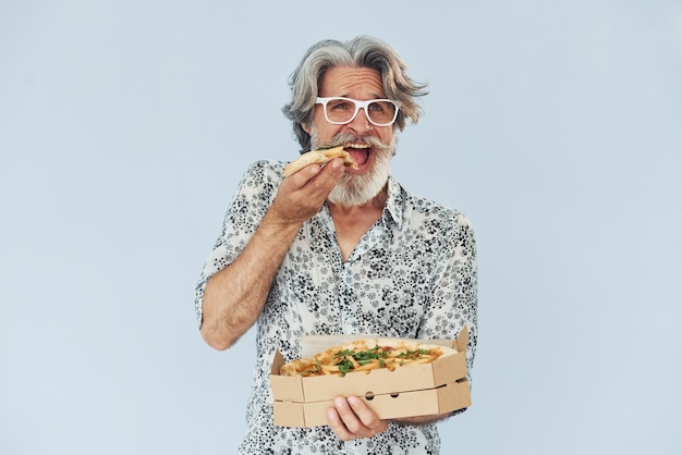Un homme moderne et élégant aux cheveux gris et à la barbe à l'intérieur mange de la pizza