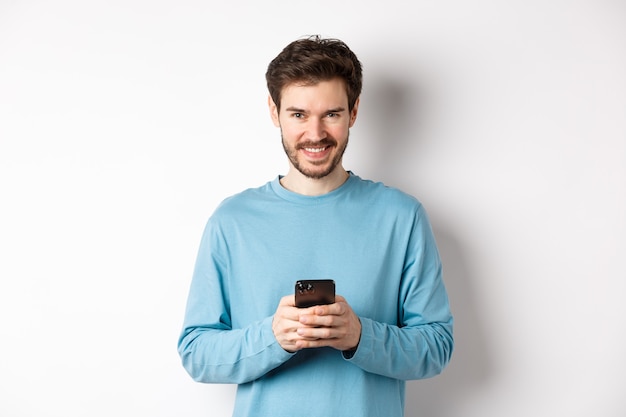 Homme moderne souriant à l'aide de smartphone et à la recherche de plaisir à la caméra. Guy en sweat-shirt avec téléphone portable sur fond blanc.