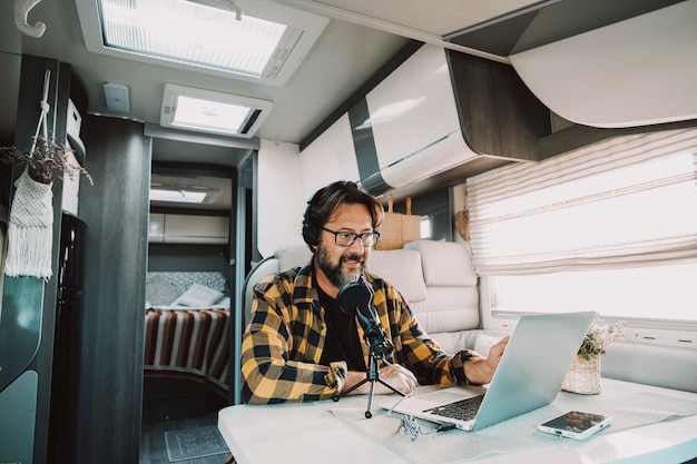 Un homme moderne travaille avec un microphone et un ordinateur portable pour enregistrer un podcast à l'intérieur d'une maison de camping-car et d'un bureau alternatif Concept de personnes et de style de vie nomade numérique Travail de voyage partout connecté