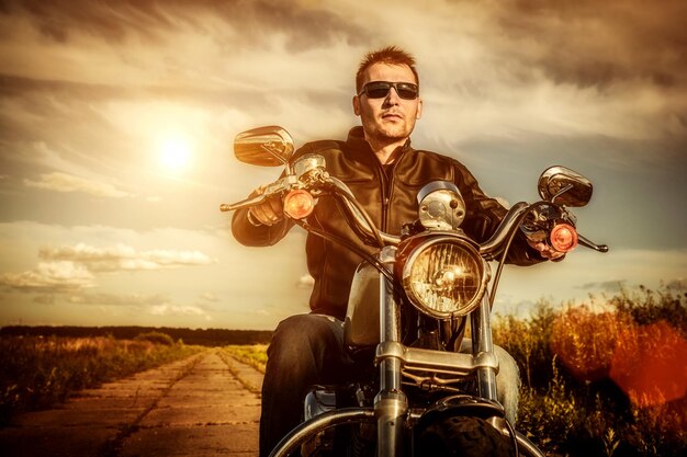 Homme de motard portant une veste en cuir et des lunettes de soleil assis sur sa moto en regardant le coucher de soleil.