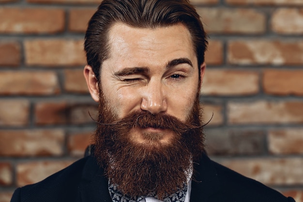 homme avec une moustache et une barbe posant contre un mur de briques et un clin de œil.