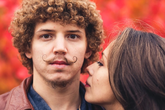 Homme moustachu aux cheveux bouclés et femme aux cheveux bruns étreignant en automne sur fond d'arbres rouges