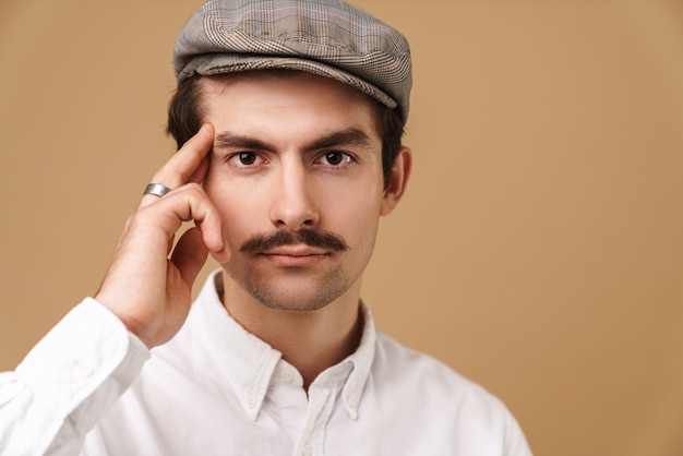 homme moustachu confiant portant un chapeau regardant à l'avant et touchant sa tempe isolée sur beige