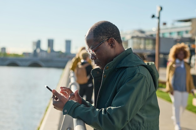 Homme mûr africain dans des lunettes tapant un message sur son téléphone portable se tenant dehors dans la ville