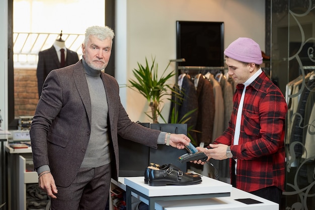 Un homme mûr aux cheveux gris et au physique sportif applique une carte de crédit à un terminal de point de vente dans un magasin de vêtements. Un client masculin avec une barbe et un vendeur dans une boutique.