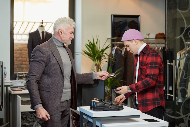 Un homme mûr aux cheveux gris et au physique sportif remet une carte de crédit à un vendeur pour payer un achat dans un magasin de vêtements. Un client masculin avec une barbe et un vendeur dans une boutique.