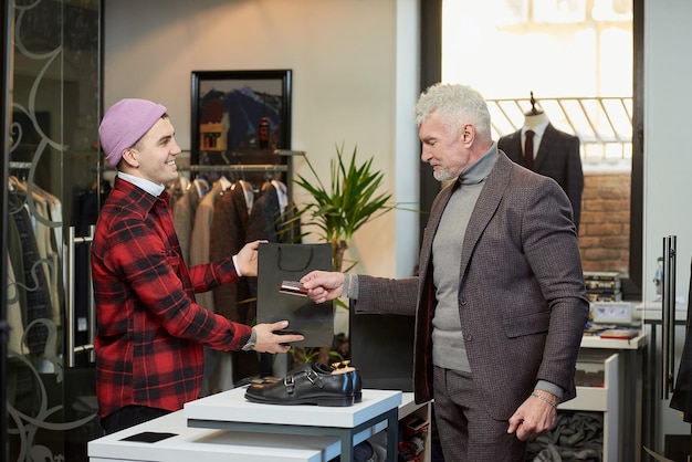 Un homme mûr aux cheveux gris et au physique sportif remet une carte de crédit à un vendeur pour payer un achat dans un magasin de vêtements Un vendeur souriant donne un sac en papier à un client masculin