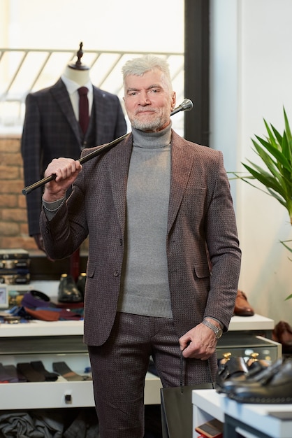 Un homme mûr aux cheveux gris et au physique sportif tient deux sacs en papier noir avec des achats et une canne en fibre de carbone sur son épaule dans un magasin de vêtements. Un client avec une barbe dans une boutique