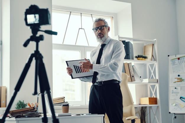 Homme mûr confiant en chemise élégante et cravate montrant un graphique et partageant une expérience commerciale tout en réalisant une vidéo sur les réseaux sociaux