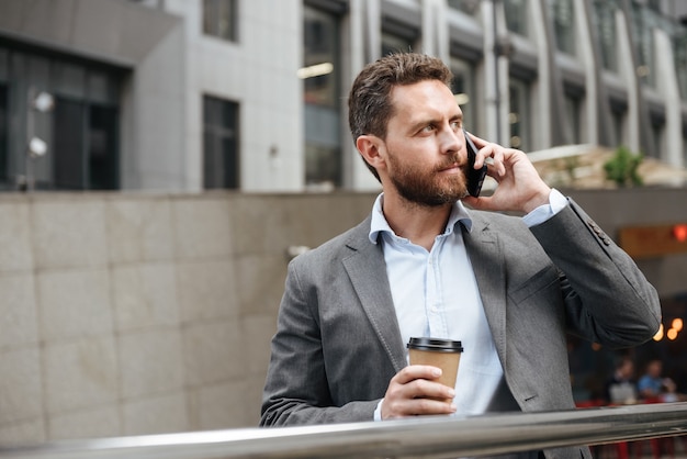 Homme mûr en costume gris à côté, en position debout et tenant du café à emporter en face du centre d'affaires moderne pendant l'appel d'affaires