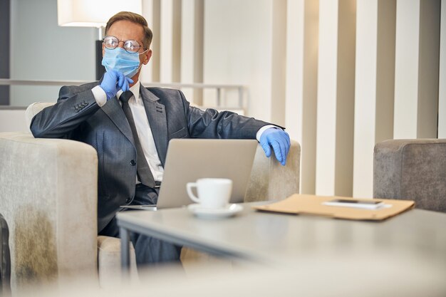 Homme mûr élégant en dlasses portant des gants de protection et un masque tout en utilisant un ordinateur portable et en buvant du café avant le vol