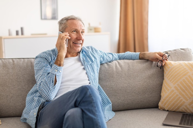 Homme mûr gai parlant au téléphone portable se reposant sur le divan