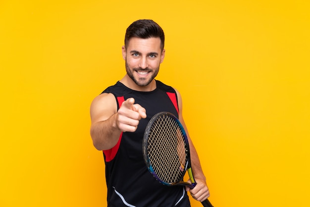 Homme sur mur jaune isolé jouant au tennis et pointant vers l'avant