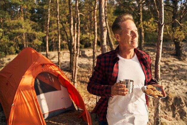 Un homme mûr joyeux se tient dans la forêt près de la tente avec une tasse de café et une assiette de biscuits