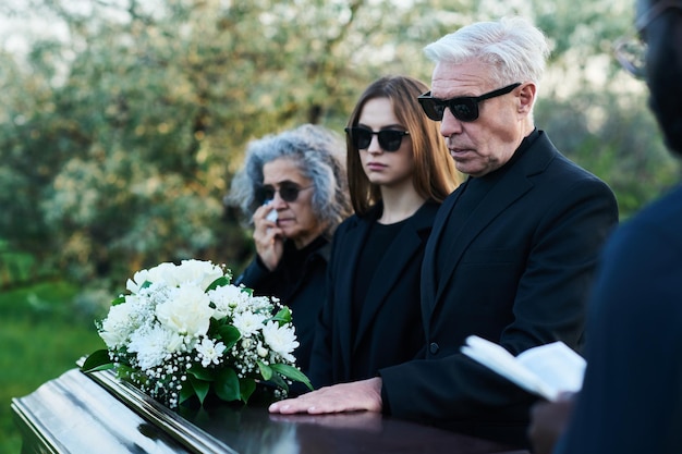 Photo homme mûr en lunettes de soleil et tenue noire gardant la main sur le couvercle du cercueil fermé