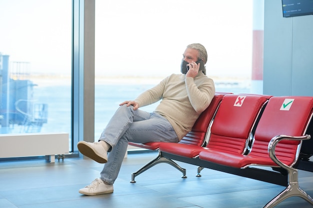 Homme mûr en masque assis sur une chaise et parlant au téléphone portable dans la salle d'attente de l'aéroport pendant la pandémie