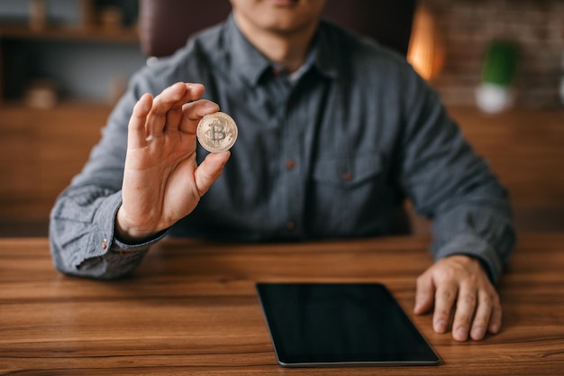 Un homme mûr montre une pièce de monnaie bitcoin à table avec une tablette avec un écran vide à l'intérieur du bureau recadré en gros plan