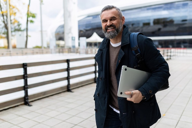 Homme mûr avec ordinateur portable et sac à dos se dépêchant de travailler en souriant regardant la caméra