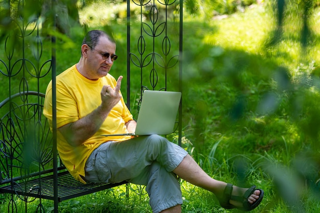 Homme mûr avec un ordinateur portable travaillant à l'extérieur dans le jardin et montrant un putain de geste dans le moniteur