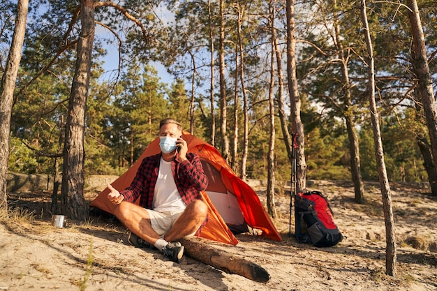 Un homme mûr porte un masque médical alors qu'il est assis près d'une tente dans la nature et parle au téléphone portable