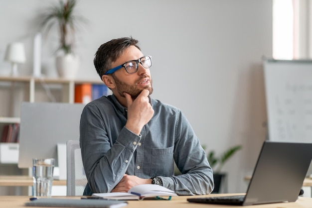 Homme mûr réfléchi assis au bureau et regardant de côté travaillant avec un ordinateur portable à la recherche de créativité