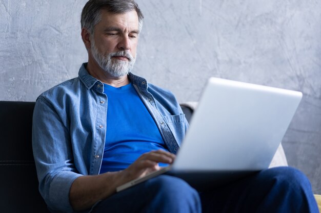 Homme mûr souriant assis sur un canapé en train de taper sur un ordinateur portable dans un salon. Travail à domicile pendant le confinement. Distanciation sociale Auto-isolement