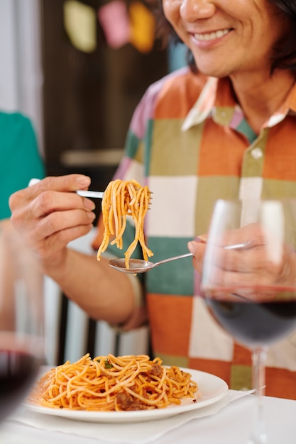 Homme mûr souriant mangeant des spaghettis bolognaise avec une cuillère et une fourchette au dîner à la maison