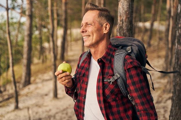 Un homme mûr souriant marche avec un sac à dos dans la forêt et a une pomme verte fraîche sur le chemin