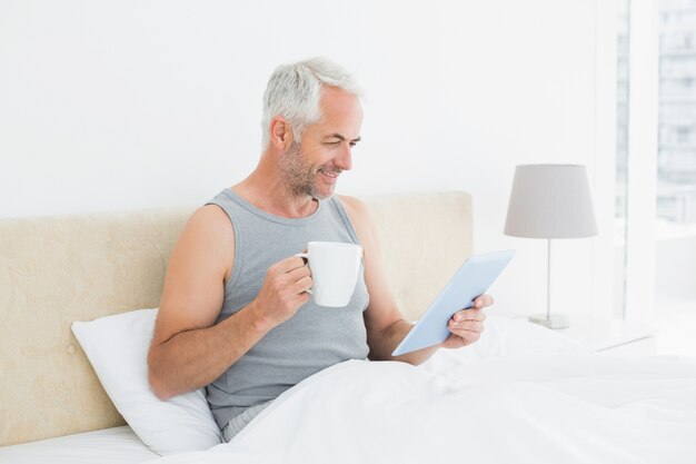 Homme mûr souriant avec tablette numérique et table basse au lit