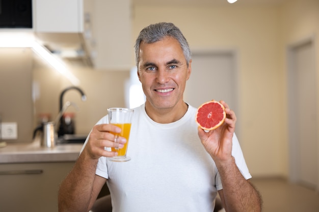 Homme mûr en t-shirt décontracté blanc tenant la moitié du pamplemousse et un verre de jus d'agrumes et souriant