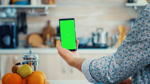 Homme mûr tenant un smartphone avec écran vert pendant le petit-déjeuner dans la cuisine. Personne âgée avec chroma key maquette isolée pour un remplacement facile