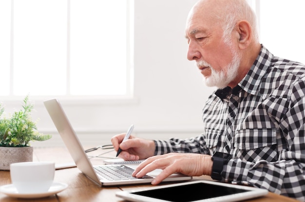 Homme mûr utilisant un ordinateur portable et écrivant dans le bloc-notes au bureau à domicile. Éducation, concept de recette