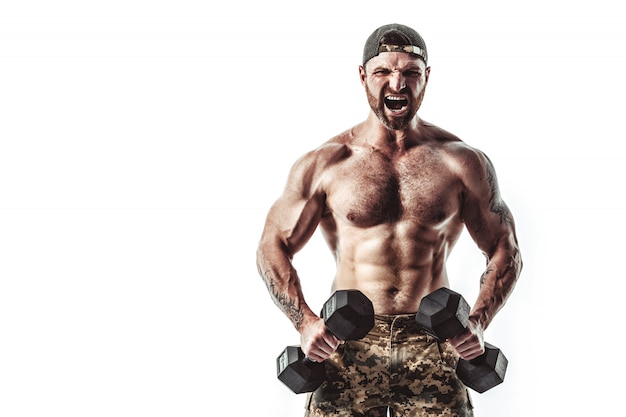 Homme musclé athlète bodybuilder en pantalon de camouflage avec un entraînement de torse nu avec haltère sur un mur blanc.